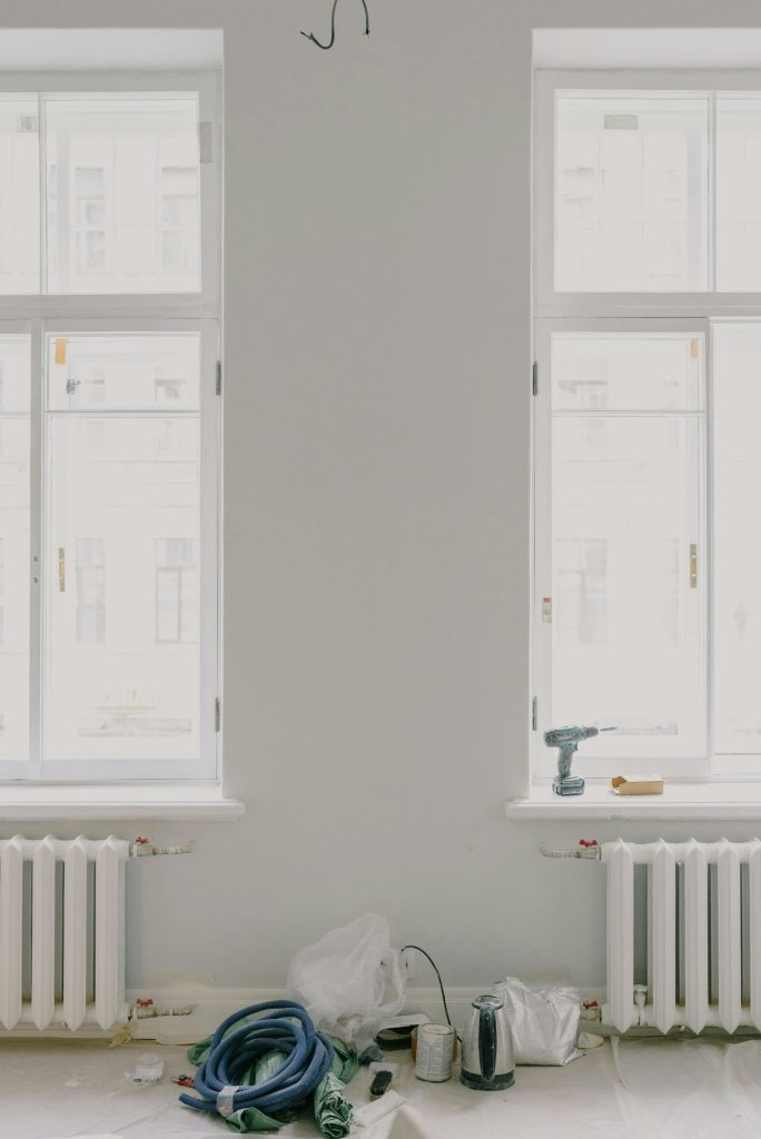 Interior of light room with white walls and new wooden windows above radiators during renovation with repairing tools and teapot located on floor